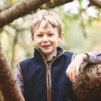 Smiling boy wearing navy gilet leaning on tree